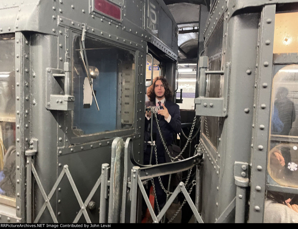 A guy wearing a suit standing on the outside part of the vestibule between the two Arnine Cars